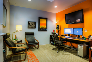 Furnished office in Creative Bloc building. Desk with computer monitors on it. Two black chairs lining the walls.