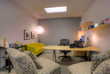 Furnished office in Creative Bloc building. Desk with computer on it. Brown chairs opposite the desk.